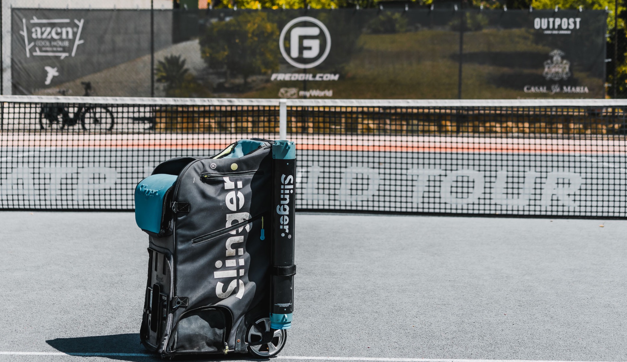 Slinger ball machine on hard court with FREDGIL sponsor banners on the net